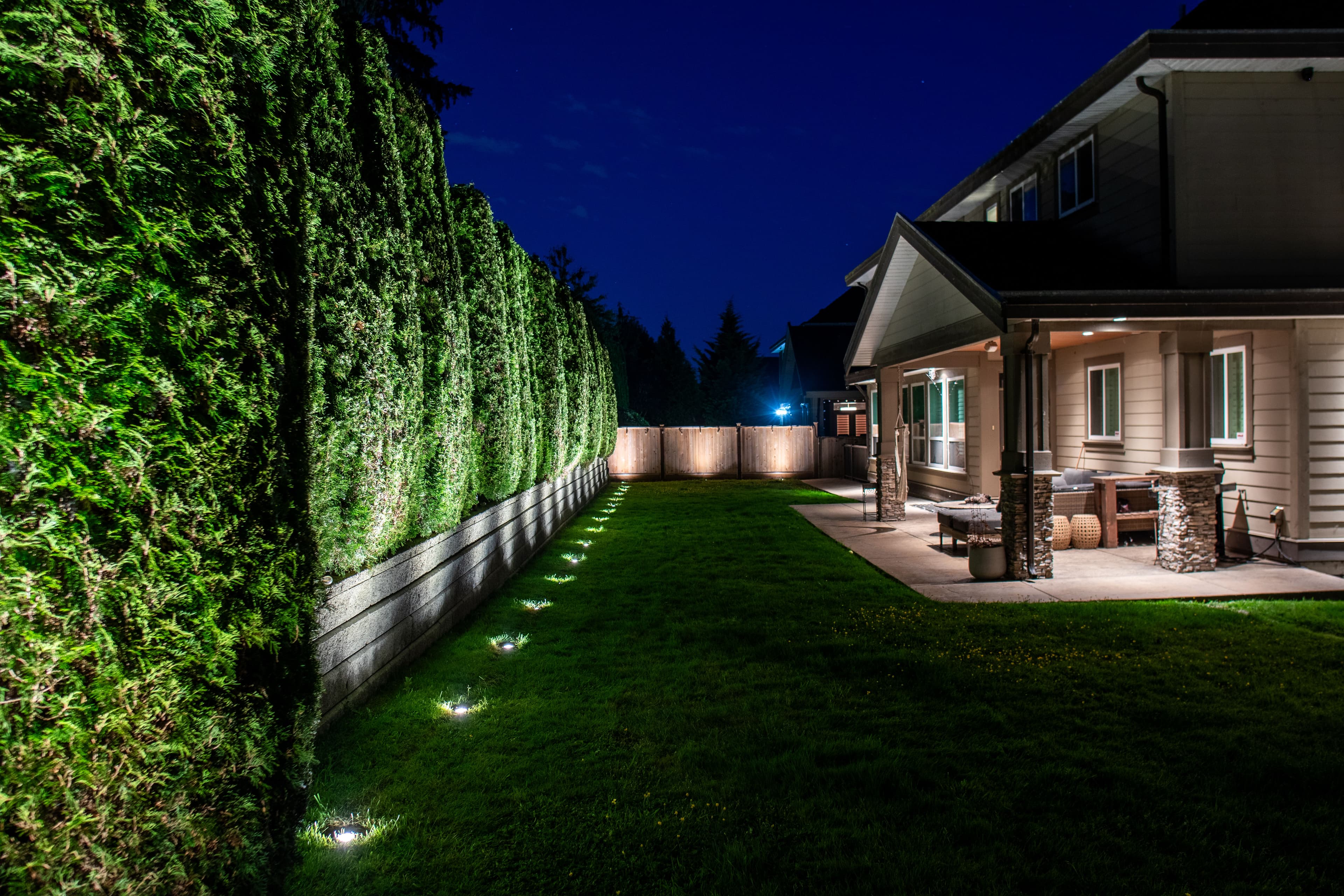 Elegant backyard with illuminated hedges, green lawn, and outdoor seating at night.